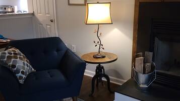 A living room with a blue armchair, a small round table with a lamp, a fireplace, and a kitchen in the background.