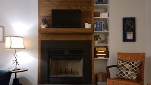 A living room with a fireplace, a television mounted above it, a bookshelf, and a chair with a patterned cushion. - New construction! 1 BD/2 BA Top of Wintergreen mountain. Sleeps 4, pet-friendly (Roseland)