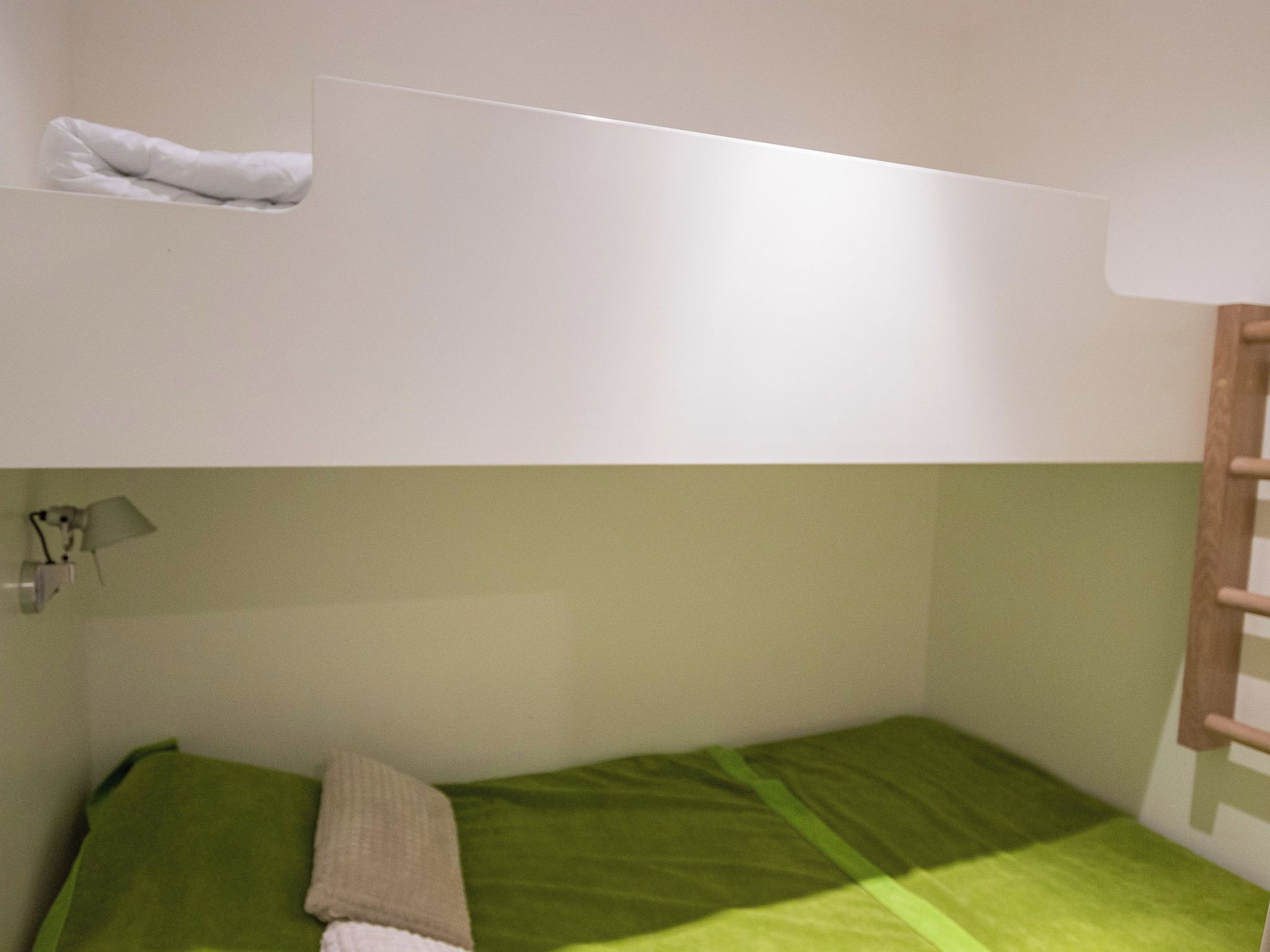 A bunk bed with a green mattress and a pillow, a small shelf with a towel, and a wall-mounted lamp.
