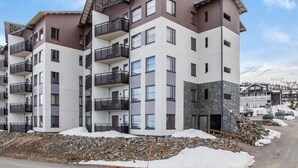 Un immeuble résidentiel à plusieurs étages, doté de balcons, d’une façade en pierre et offrant une vue sur une montagne enneigée.
