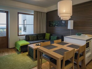 Una sala moderna con un sofá, una mesa de comedor con sillas, una zona de cocina con microondas y un amplio ventanal con cortinas.
