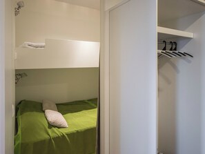 Una cama litera con un colchón verde y una almohada blanca, una pared blanca y un perchero con ropa colgada.