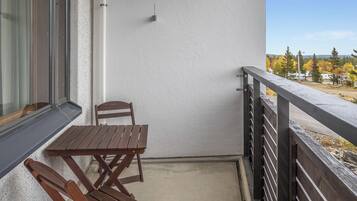 Un balcon avec une table et des chaises en bois, une fenêtre et une vue sur des arbres et un véhicule.