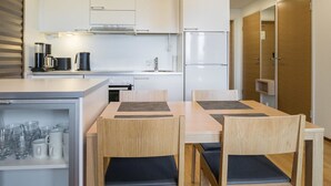 Une cuisine moderne avec des armoires blanches, un four encastré et un réfrigérateur. On y trouve un coin repas avec une table et des chaises en bois, ainsi qu’une étagère avec des verres et des assiettes.