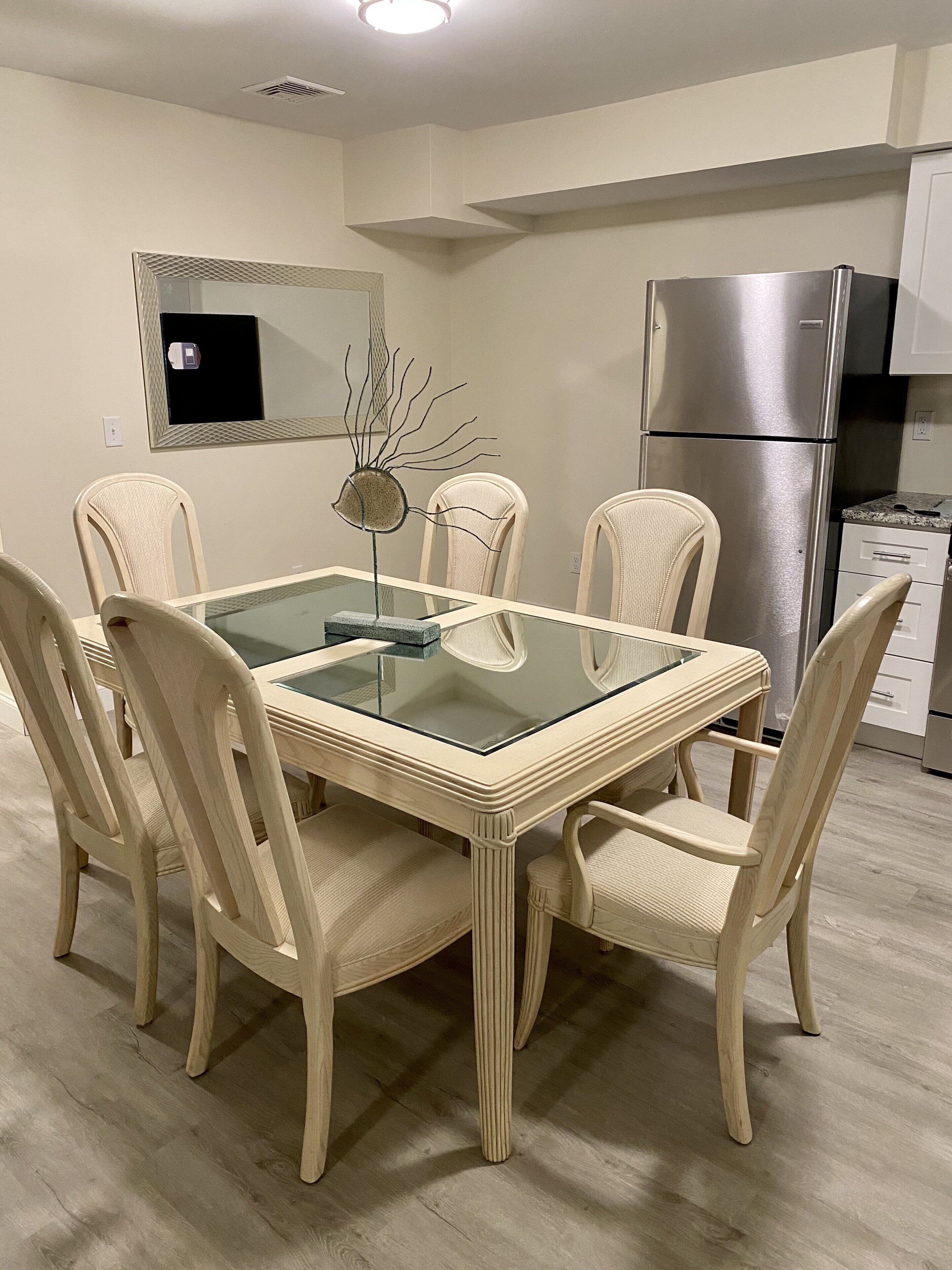 A dining area with a glass-top table surrounded by four chairs, a modern kitchen with a stainless steel refrigerator, and a wall-mounted flat-screen TV.