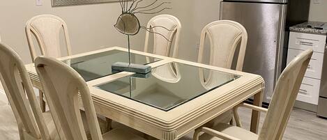 A dining area with a glass-top table surrounded by four chairs, a modern kitchen with a stainless steel refrigerator, and a wall-mounted flat-screen TV.