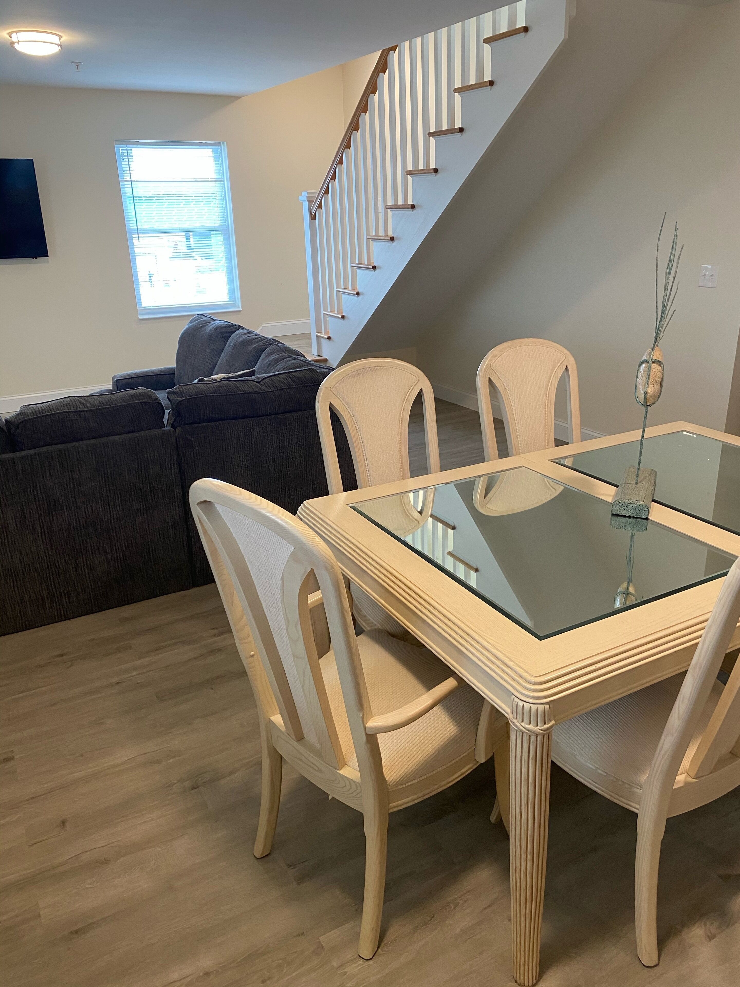 Un coin repas avec une table à plateau de verre et quatre chaises, un escalier en arrière-plan et un canapé dans le salon.