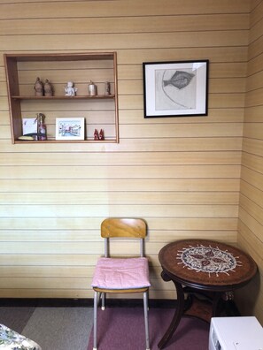 A room with a wooden paneled wall, a small wooden shelf with decorative items, a framed fish illustration, a chair with a pink cushion, and a round wooden table with a patterned tablecloth.