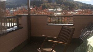 A balcony with a wooden chair, a table with a floral tablecloth, and a clear view of a mountain and buildings in the background. - Minshuku Tomato House (Tamura)