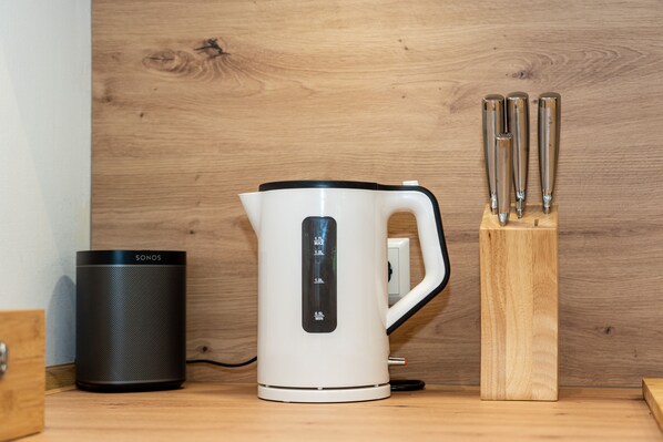 Une bouilloire électrique blanche avec une base noire, un haut-parleur SONOS noir et un bloc à couteaux en bois contenant trois couteaux, le tout posé sur une surface en bois.