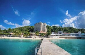 A coastal hotel with a swimming pool, beach, and a pier leading to the water.