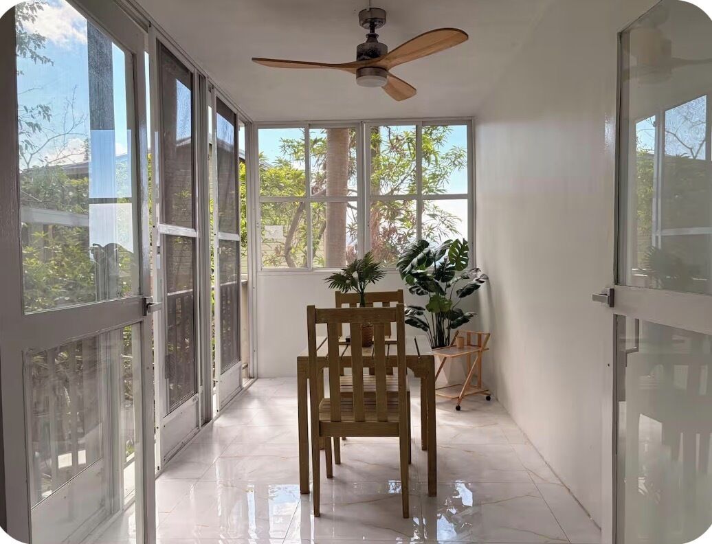 A sunlit room with a ceiling fan, a dining table with chairs, and large windows with a view of trees.