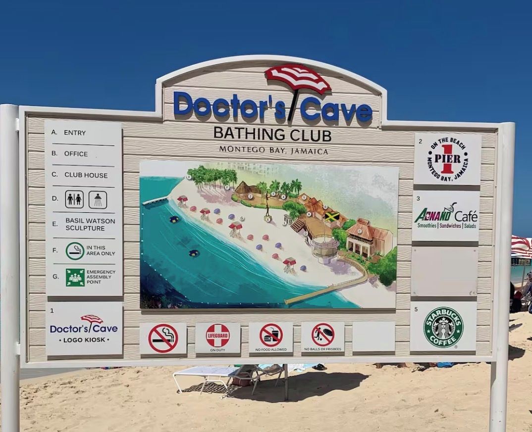 A sign for Doctor's Cave Bathing Club with a beach scene illustration, a Pier 1 logo, and nearby amenities like an Awani Cafe and Starbucks.