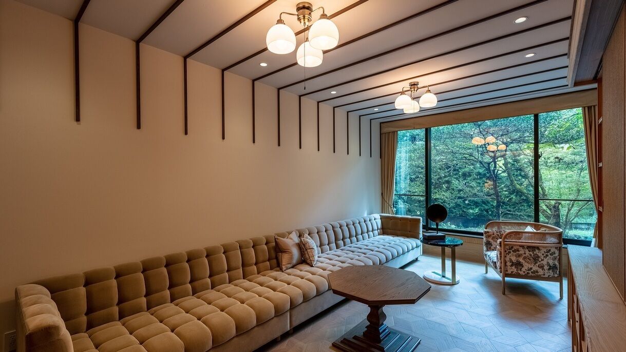 Un salon moderne avec un canapé, une table basse ronde et un fauteuil. De grandes fenêtres offrent une vue sur les arbres.