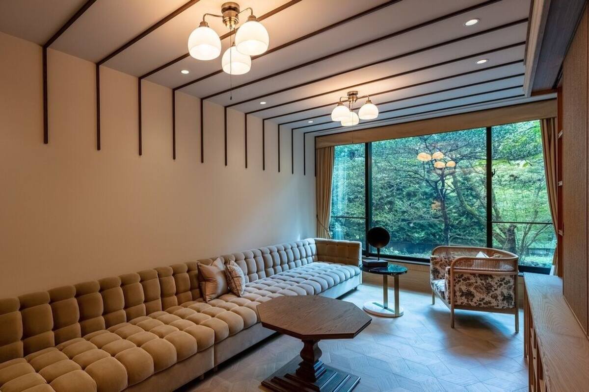 A modern living room with a sofa, a round coffee table, and a chair. There are large windows with a view of trees.