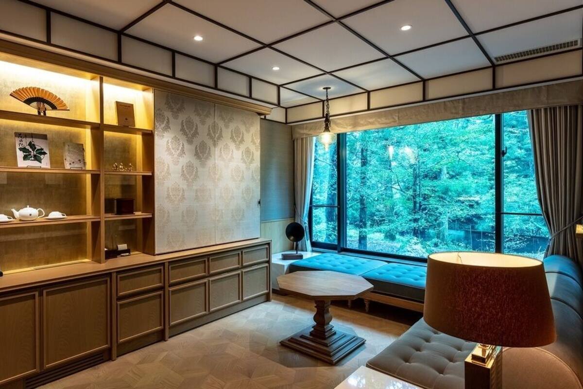 A room with a large window, a wooden cabinet with shelves, a round table, and a lamp.