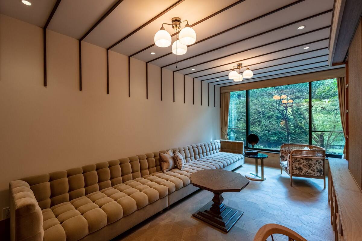 A modern living room with a large sofa, a coffee table, and a view of a forest through large windows.