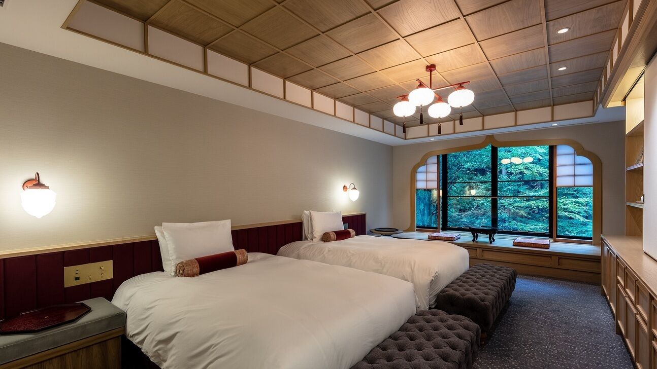 Une chambre d’hôtel avec deux lits, un banc, un bureau et une grande fenêtre donnant sur des arbres.