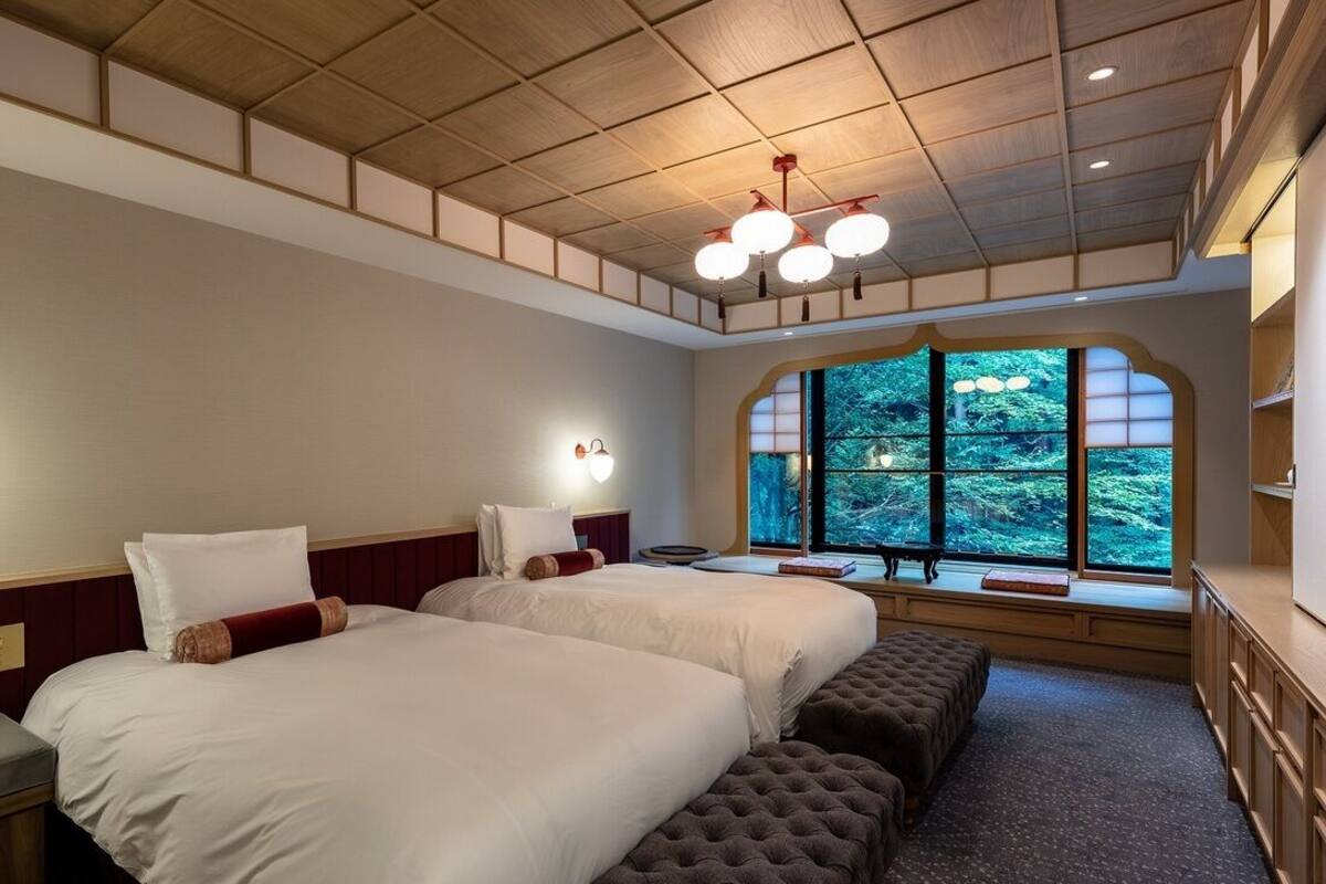 A hotel room with two beds, a bench, a desk, and a large window with a view of trees.