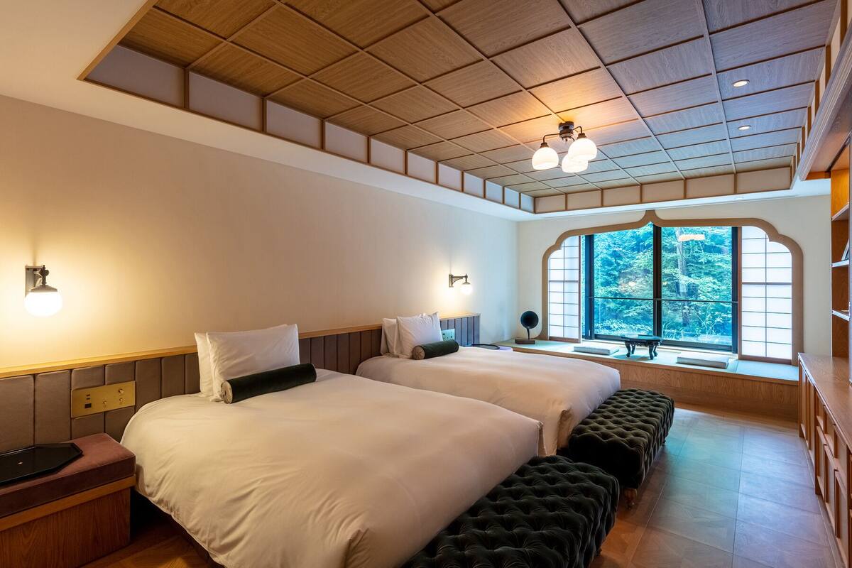 A hotel room with two beds, a large window with a view of trees, a wooden ceiling, and a bench by the window.
