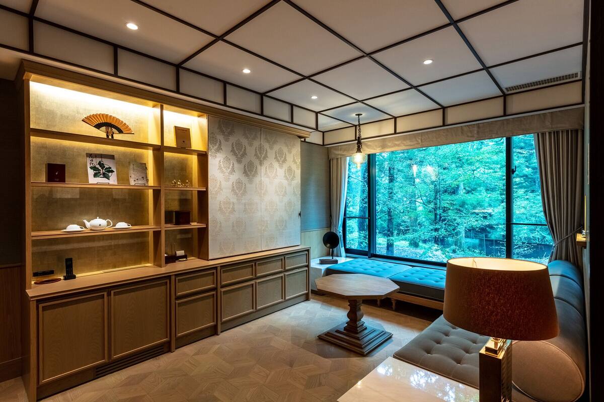 A room with a large window, a wooden cabinet with tea sets, and a lamp.