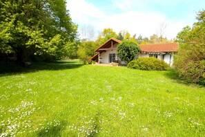 A house with a red-tiled roof, surrounded by a lush green lawn with white flowers, and a large tree to the left.