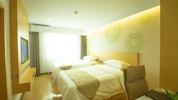 A hotel room with a large bed, a desk, and a chair. There is a window with curtains, a wall-mounted air conditioner, and a wall with circular patterns.