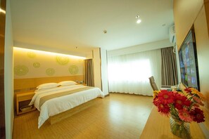 Une chambre d’hôtel avec un lit, un bureau et une télévision. Il y a une fenêtre avec des rideaux et un vase rempli de fleurs sur le bureau.