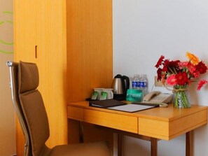 Ein Hotelzimmer mit einem Holztisch, einem braunen Stuhl, einem Blumenstrauß, einem Telefon, einem Wasserkocher und ein paar Flaschen Wasser.