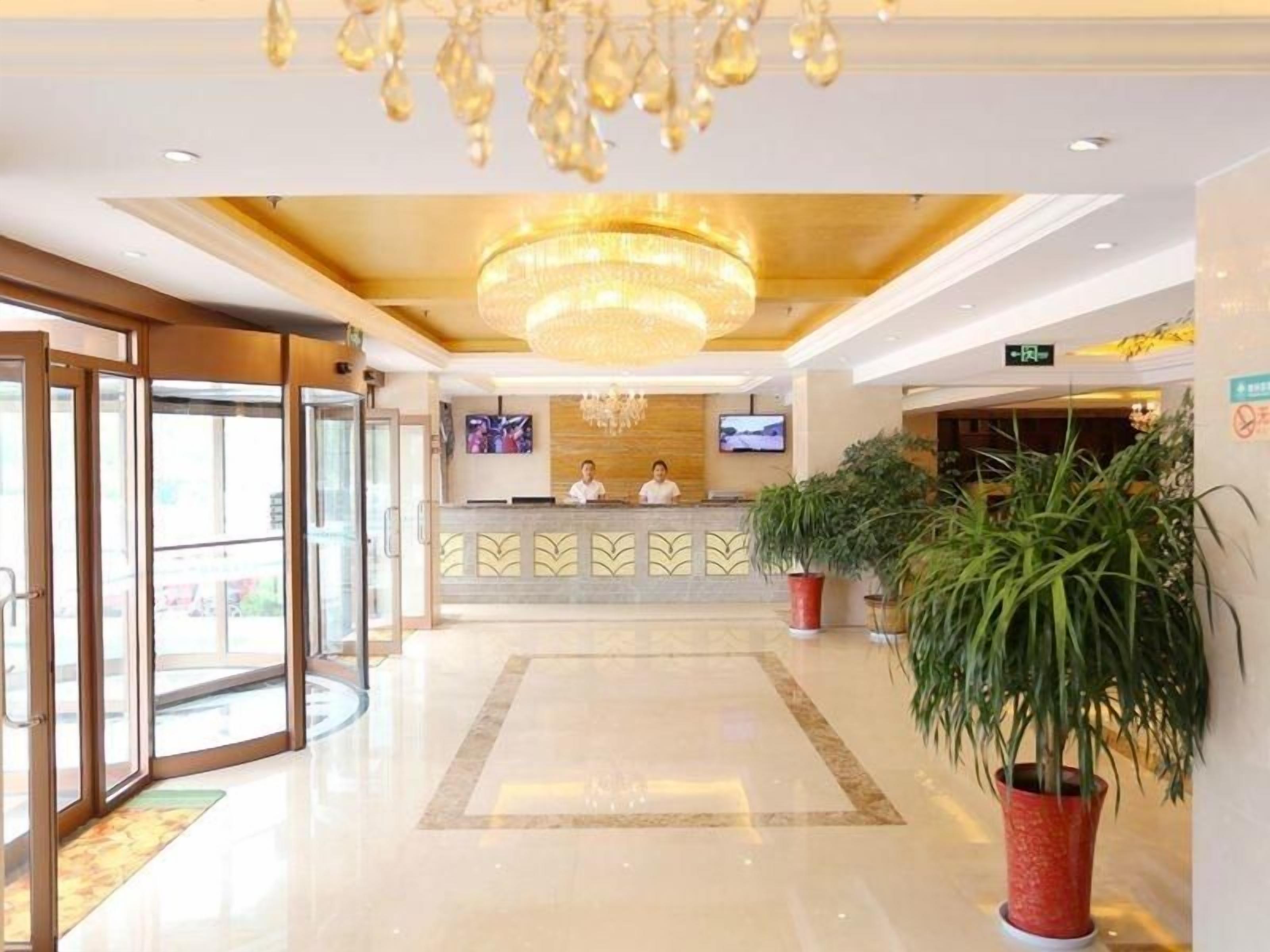 Zona de recepción de un hotel con una araña, un mostrador de recepción, dos empleados y plantas en macetas.