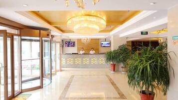 A hotel reception area with a chandelier, a reception desk, two staff members, and potted plants.