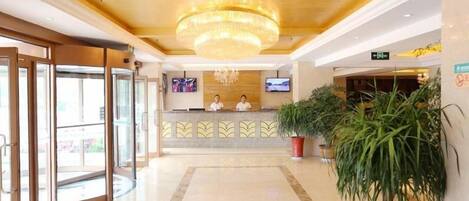 A hotel reception area with a chandelier, a reception desk, two staff members, and potted plants.