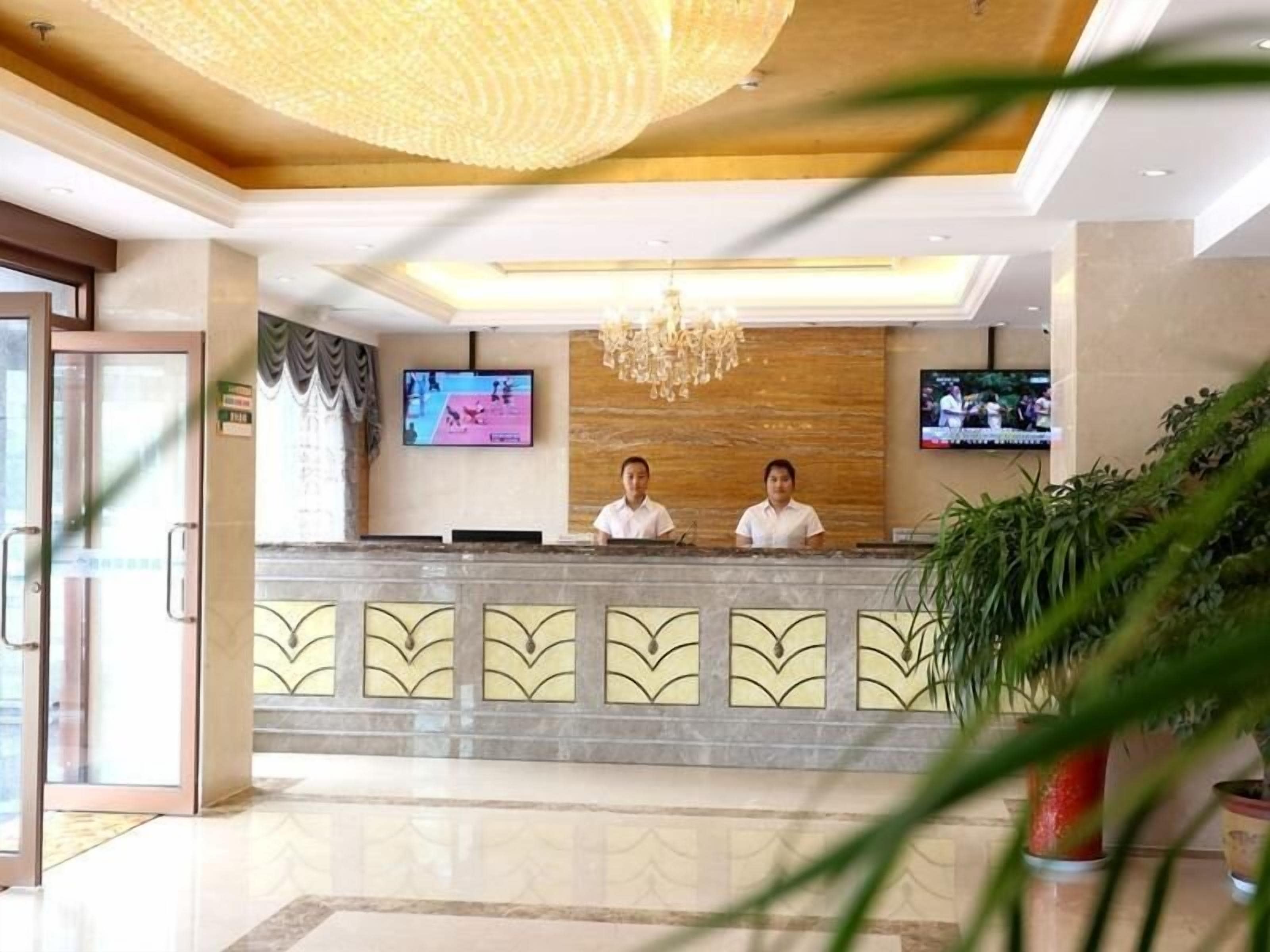A hotel reception area with a marble counter, two receptionists, a television screen, and a chandelier.