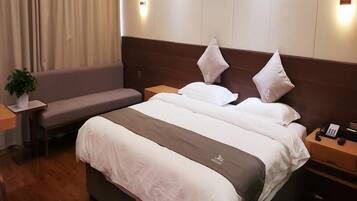 A hotel room with a bed, a bedside table, a nightstand, a lamp, a plant, a sofa, and a window with curtains.