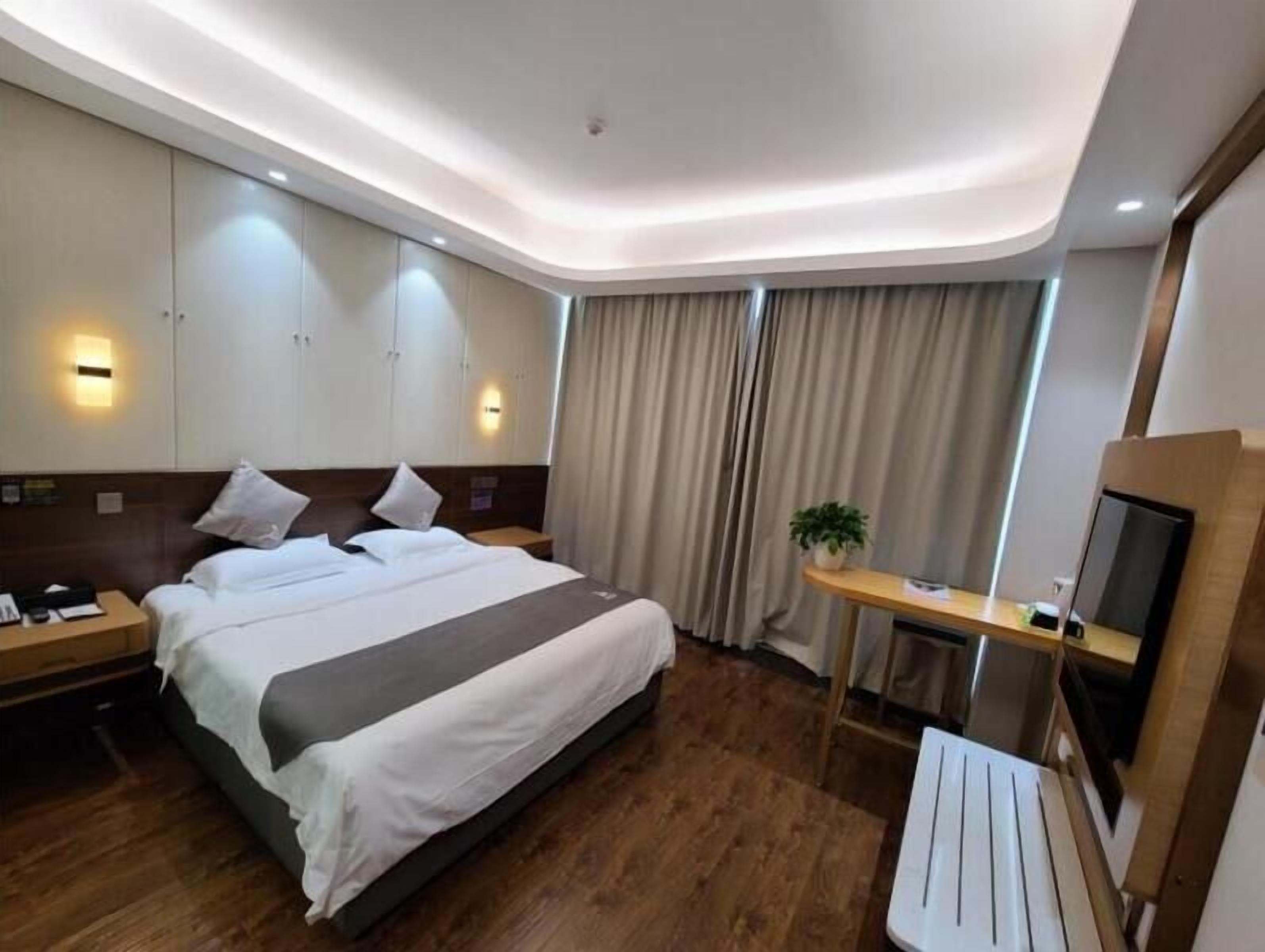 A modern hotel room with a large bed, bedside tables, a desk with a chair, a TV, and a window with curtains.