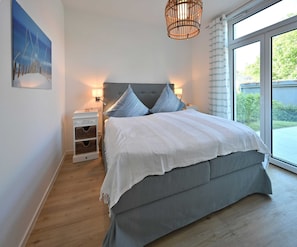 A bedroom with a large bed, two bedside tables, a nightstand, a hanging light fixture, and a view of greenery outside.
