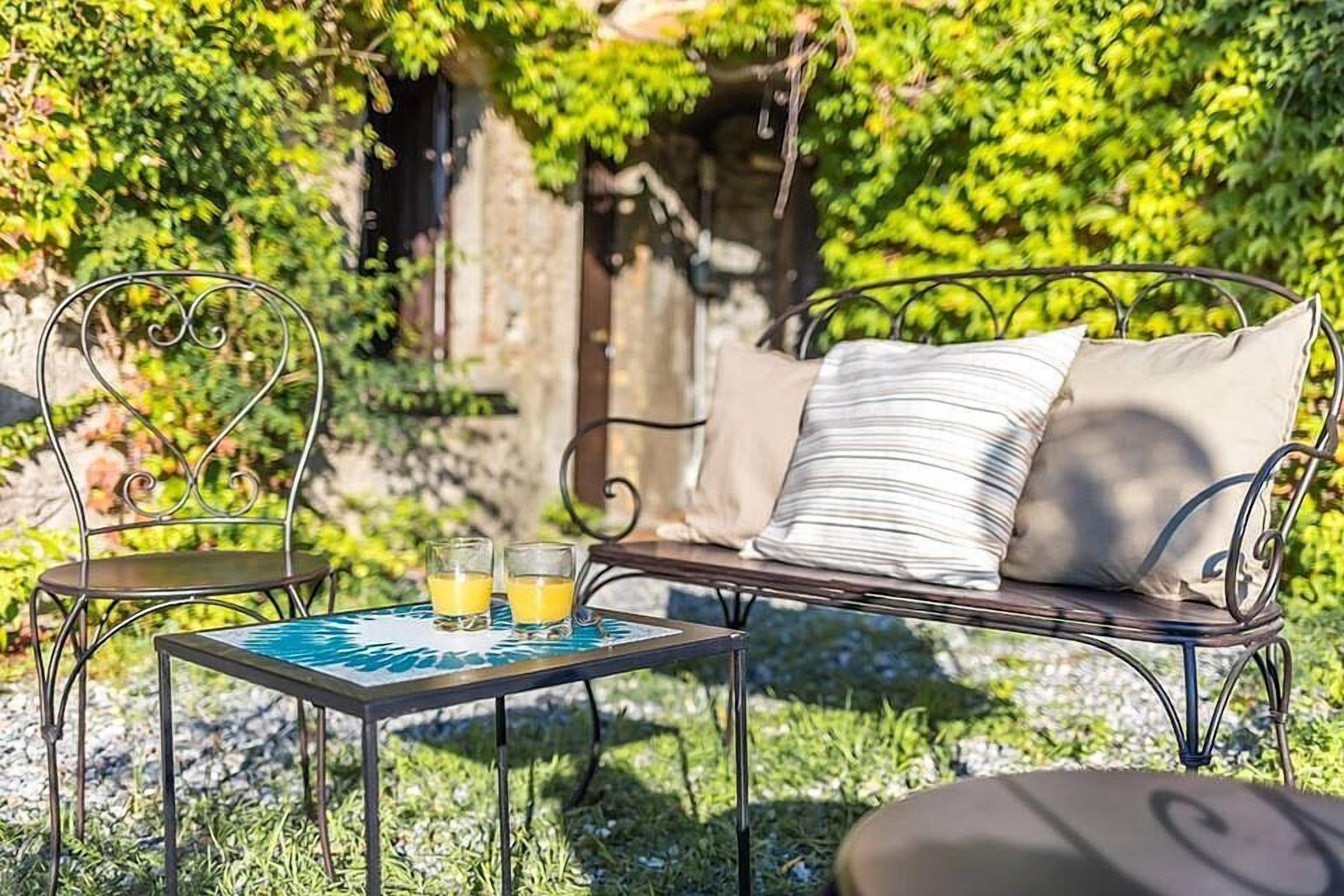An outdoor seating area with a metal table and chairs, a cushioned bench, and a glass of orange juice.