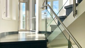 Un escalier moderne avec des rampes en verre et un sol noir, menant à un espace ouvert doté de grandes fenêtres.