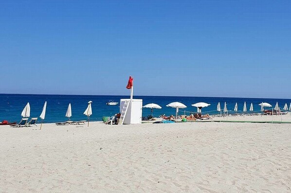 Ein Strand mit weißen Sonnenschirmen und Liegestühlen, einem Rettungsturm und dem Meer im Hintergrund.
