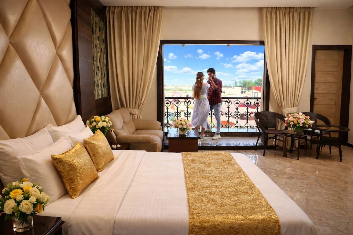 Habitación de hotel con una cama grande, un sofá, una mesa de comedor con sillas y un balcón con barandilla.