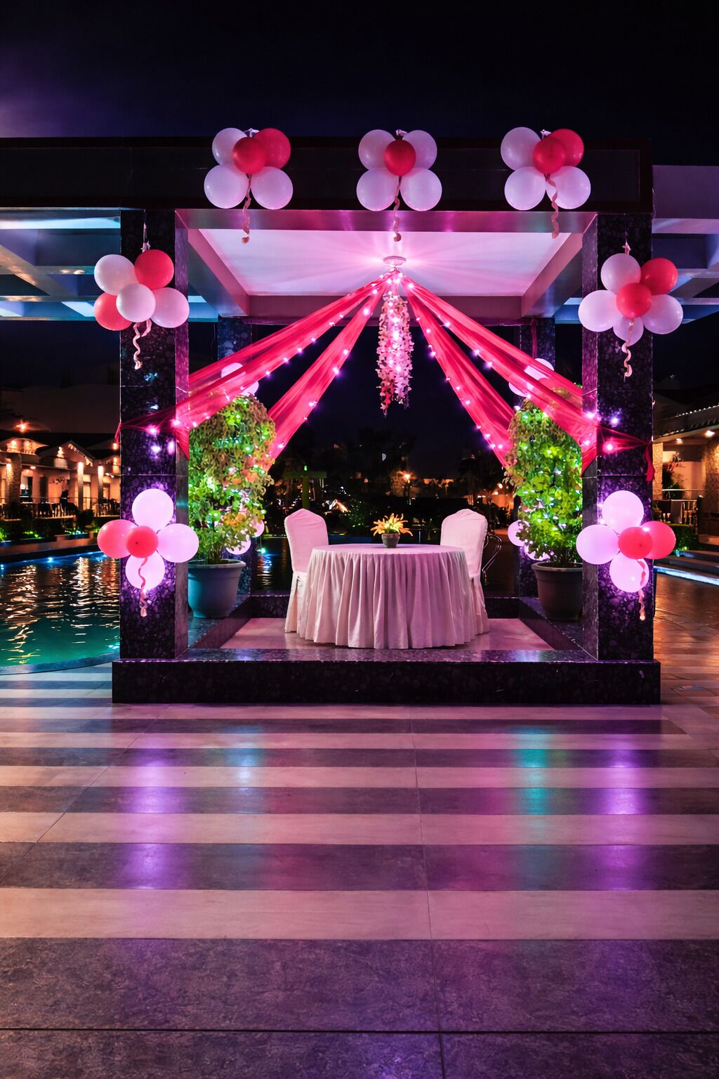 A nighttime outdoor dining area with a table and chairs, surrounded by decorative elements and a pool.