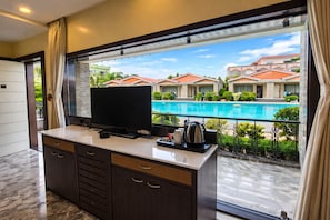 A room with a TV on a desk, a balcony with a pool view, and a stone wall. - Grand Beach Resort (Kanthi)