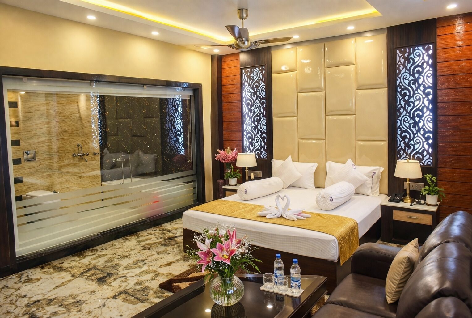 A hotel room with a large bed, a sofa, a coffee table, a vase with flowers, and a large window.
