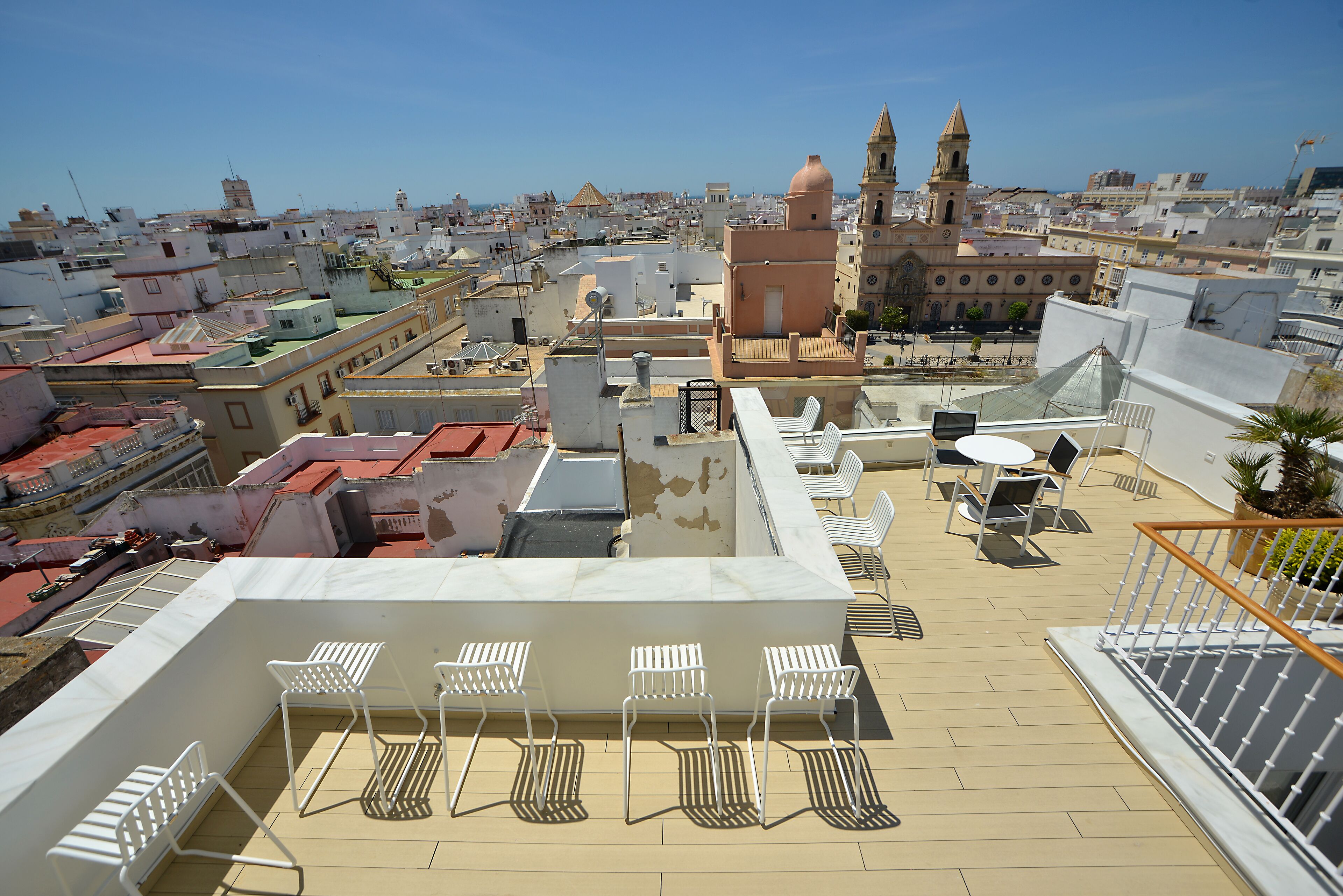 Una terraza en la azotea con sillas y una mesa blancos, con vistas a un paisaje urbano que incluye edificios históricos y una iglesia destacada.