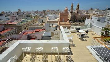 Una terraza en la azotea con sillas y una mesa blancos, con vistas a un paisaje urbano que incluye edificios históricos y una iglesia destacada.