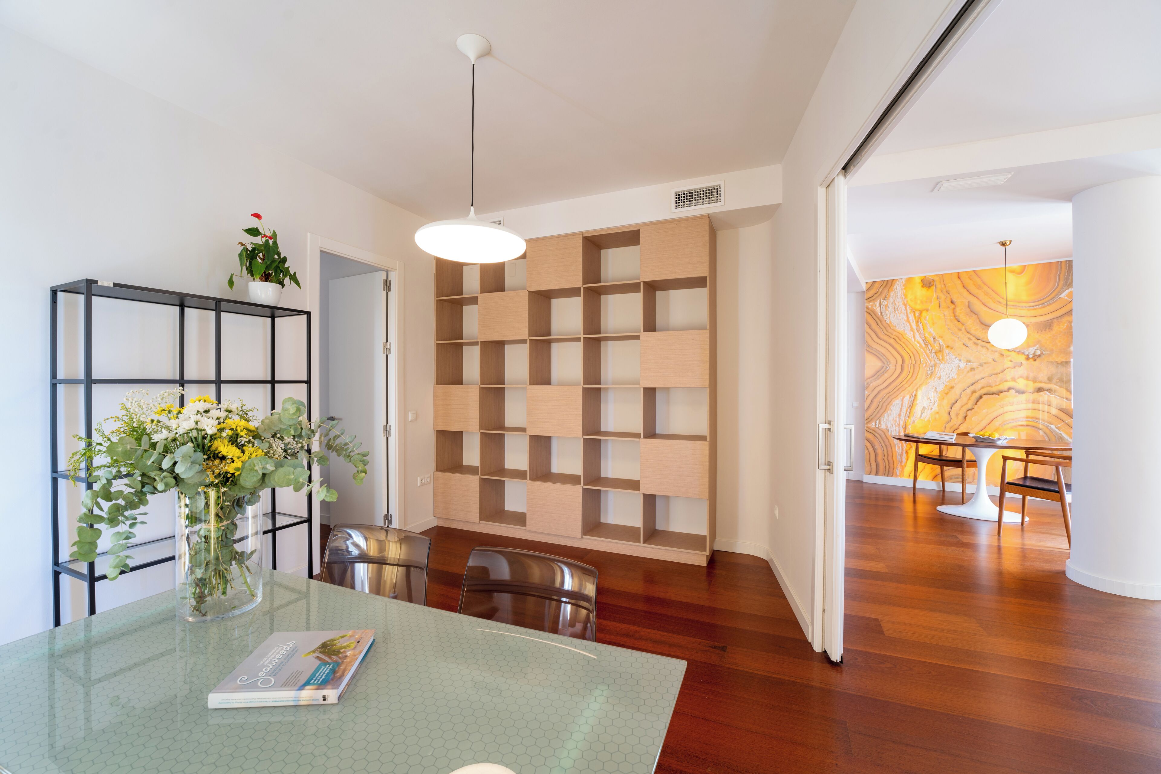A modern dining area with a glass table, metal chairs, a vase with flowers, and a wooden wall with shelves.