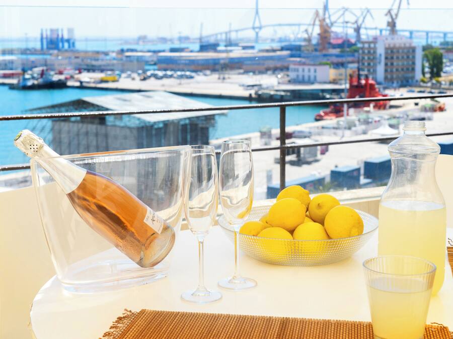 Una mesa con una botella de champán, dos copas, un cuenco de limones y una botella de limonada, con vista a un puerto y edificios al fondo.