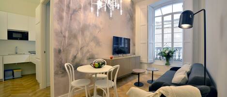 A modern living room with a sofa, a TV, a dining table with chairs, and a kitchen area with a microwave.