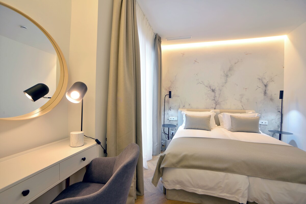 Quarto de hotel moderno com uma cama, mesas de cabeceira, uma escrivaninha com abajur e uma cadeira.