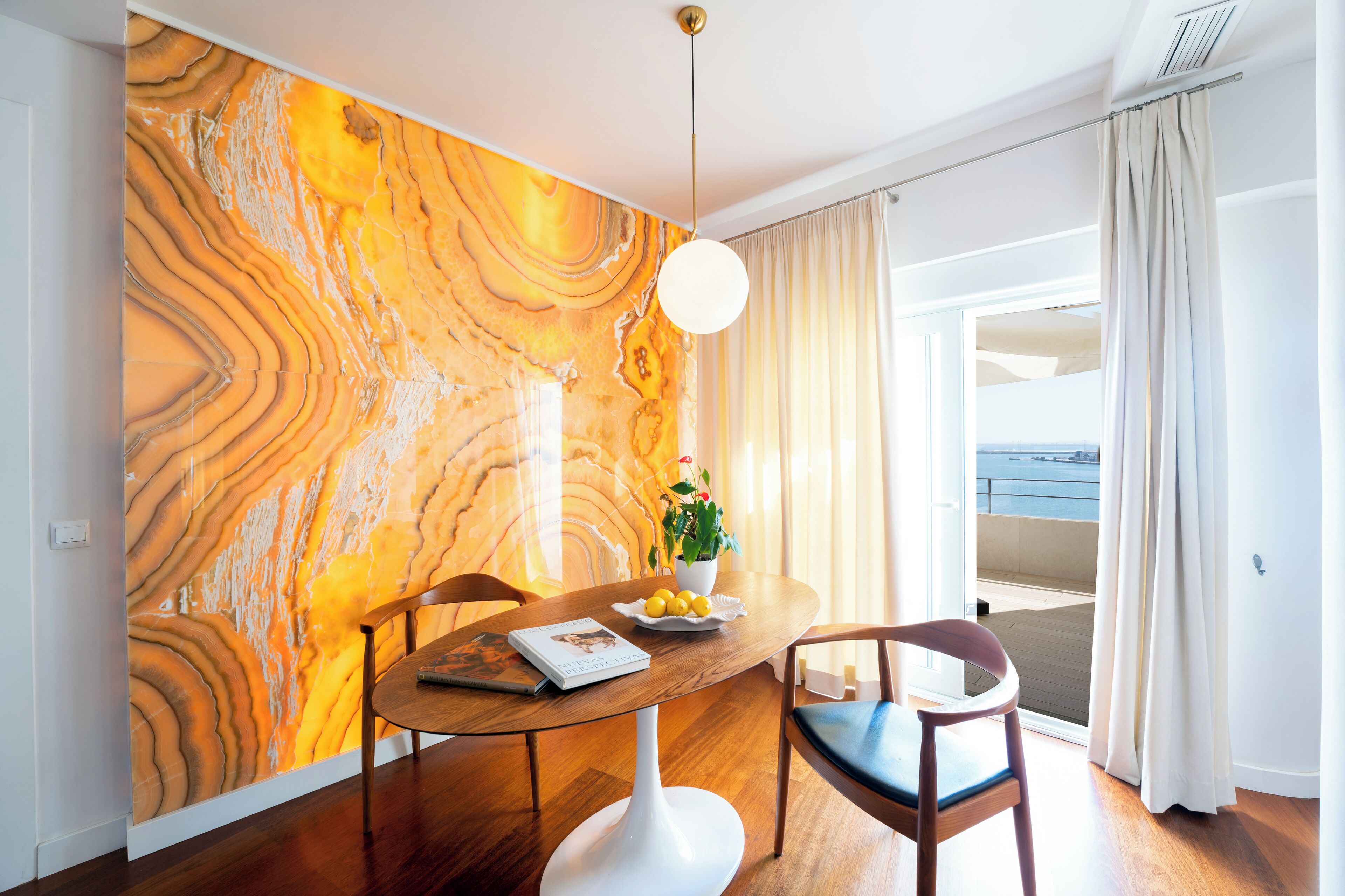 A dining area with a marble wall, a round table, two chairs, a potted plant, and a view of the sea through a balcony.
